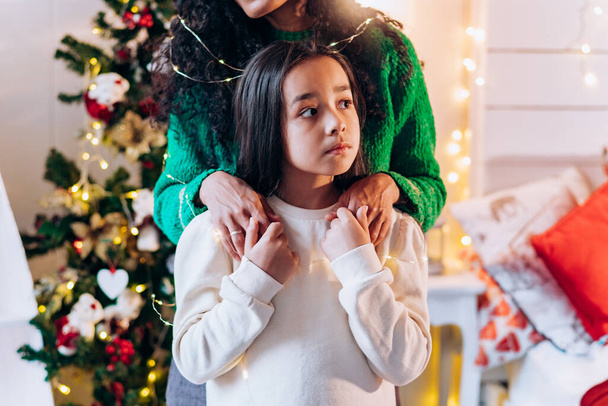 Portrait of a little girl and her mother standing behind her, putting her hands on her shoulders against the background of a Christmas tree with a garland - Photo, Image