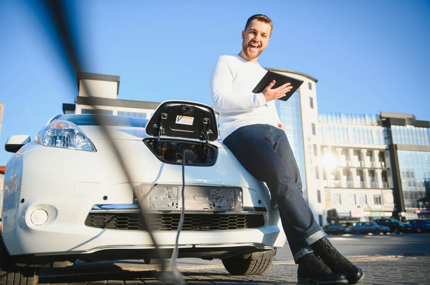 młody przystojny mężczyzna trzymający kabel ładujący w punkcie ładowania elektrycznego stojącym obok jego nowego samochodu. - Zdjęcie, obraz