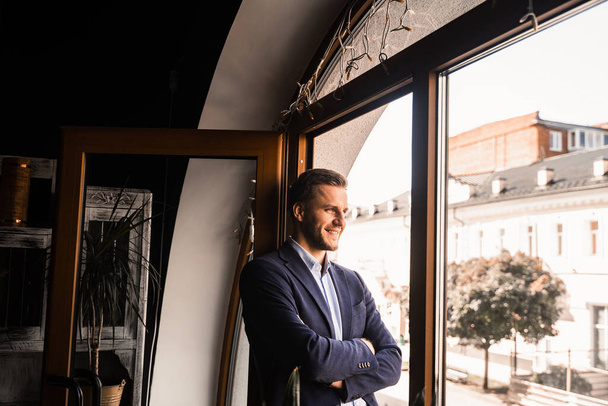 Modelo masculino guapo posando en la cafetería. hombre barbudo vestido traje de pie y sonrisa cerca de la ventana - Foto, Imagen