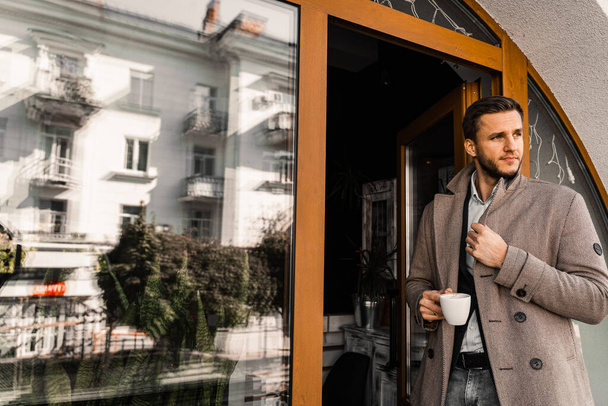 Handsome man with cup of coffee posing in coat in cafe. Male lifestyle - Foto, imagen
