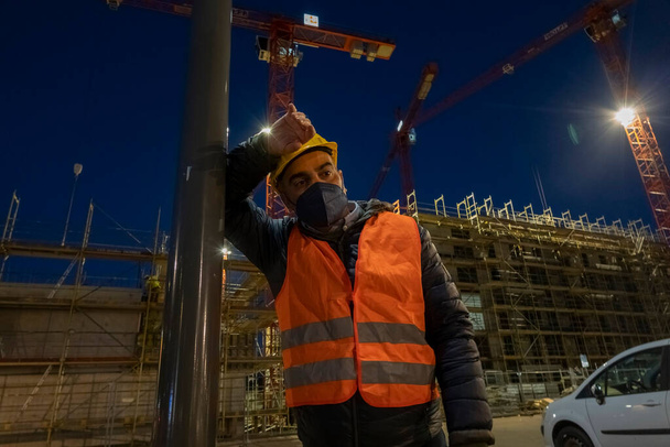 Travailleur de la construction fatigué et coûteux à l'extérieur la nuit, vêtu d'un gilet de sécurité, d'un masque médical et d'un casque de protection appuyé contre un mât lumineux - Photo, image