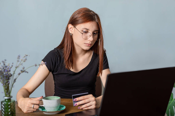 mladá žena v černém tričku s kreditní kartou v ruce se dívá na notebook, nakupuje na internetu a pije kávu. Koncepce e-commerce nebo volnočasové aktivity - Fotografie, Obrázek