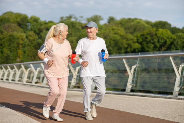Glückliches Senioren-Paar mit Flaschen läuft gemeinsam auf Fußgängerbrücke - Foto, Bild