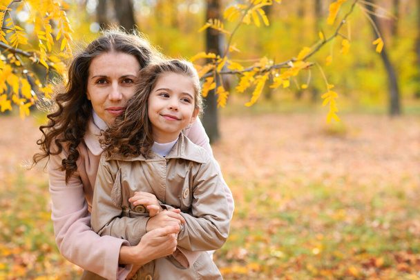Portrét matky a dcery v podzimním parku. Šťastní lidé pózují na pozadí krásných žlutých stromů. Objímají se a jsou spolu šťastní. - Fotografie, Obrázek