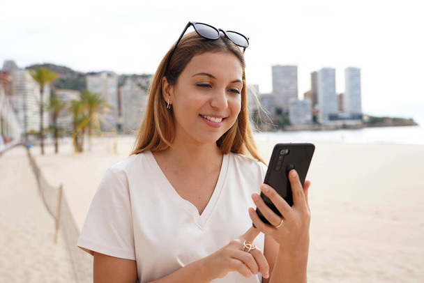 Digital nomad. Attractive millennial freelancer working reading messages during virtual chatting remotely near the beach with skyline skyscrapers on background. - Photo, Image