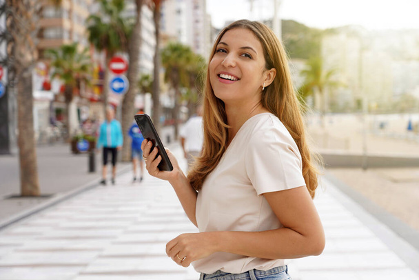 Blonde female model holding telephone looks around on promenade with blurred city background with plam trees and skyscrapers - Foto, Imagem