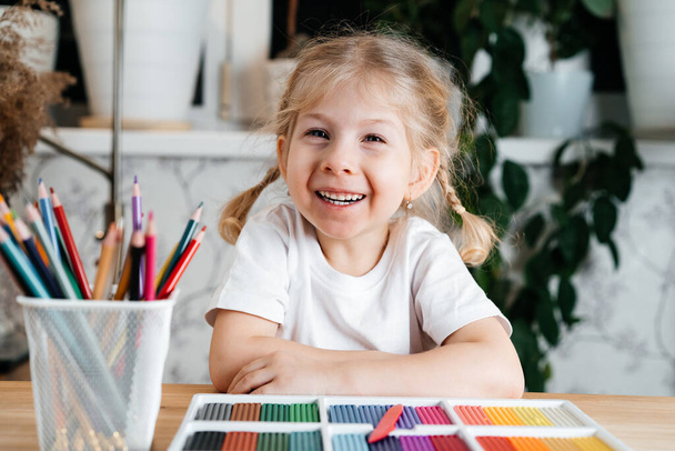 uma menina loira pequena com um sorriso encantador senta-se em uma mesa com uma paleta colorida brilhante de plasticina colorida para modelar em casa, hobbies e desenvolvimento infantil - Foto, Imagem