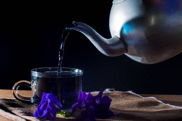 Pour Butterfly pea tea by metal pot to cup with violet flower on brown table cloth on black background. Healthy beverage for drink. Herbs and medical concept. - Fotoğraf, Görsel