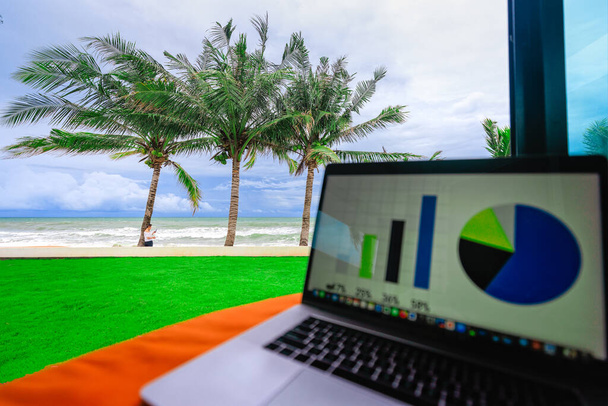 Woman holding coffee cup and smartphone standing under coconut palm tree on sea beach near beachfront house with business graph and charts on laptop blur foreground. Concept of successful life. - Photo, Image