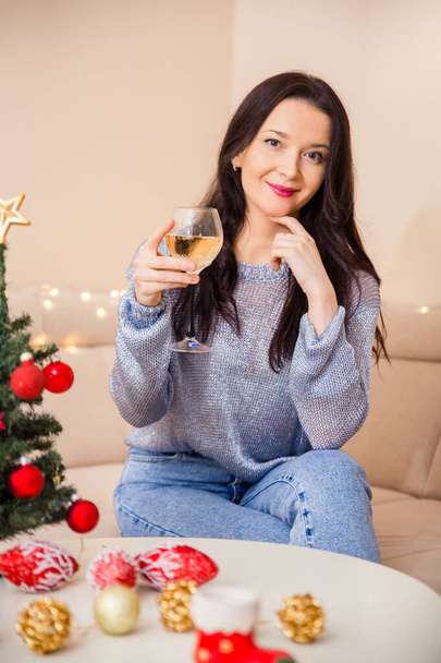 Krásná brunetka sedí v pokoji na gauči vedle vánočního stromečku zdobeného hračkami. Dívka držící v ruce sklenici vína - Fotografie, Obrázek