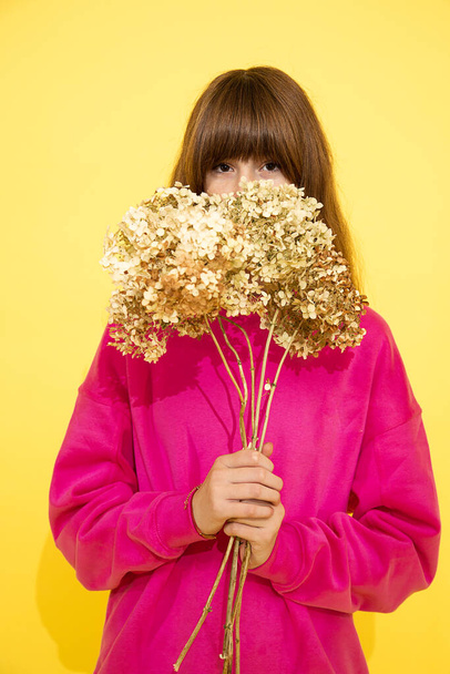 dospívající dívka s tmavými vlasy a ofinou schovaná za květem. růžový svetr, fotografie ve studiu na žlutém pozadí, portrét - Fotografie, Obrázek