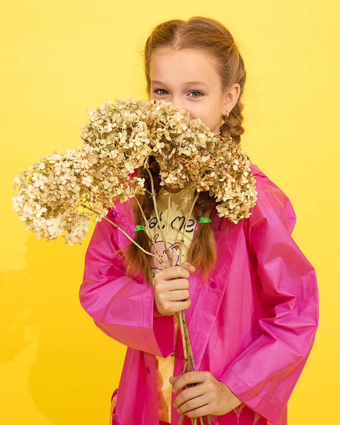 Pequena menina bonita em capa de chuva rosa com tranças escondidas atrás da flor. estúdio filmado em fundo amarelo, retrato, Dia das Mães, aniversário. foto para livro sobre crianças modernas - Foto, Imagem
