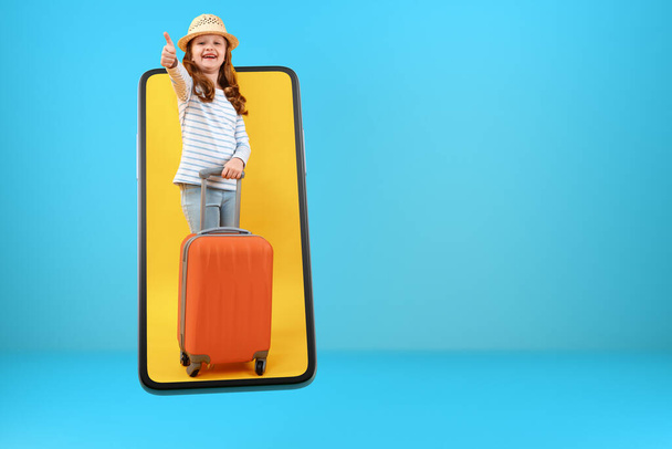 A full-length preteen girl with an orange suitcase in a smartphone. Online child on the screen of a mobile phone smiles and shows a thumb up. - Photo, Image