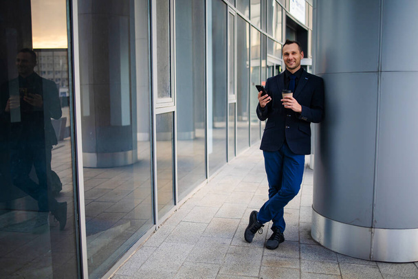 Šťastný usměvavý podnikatel v černém obleku a pomocí moderního smartphonu v blízkosti kanceláře brzy ráno, úspěšný zaměstnavatel uzavřít dohodu, stojí v blízkosti mrakodrapu kancelář - Fotografie, Obrázek