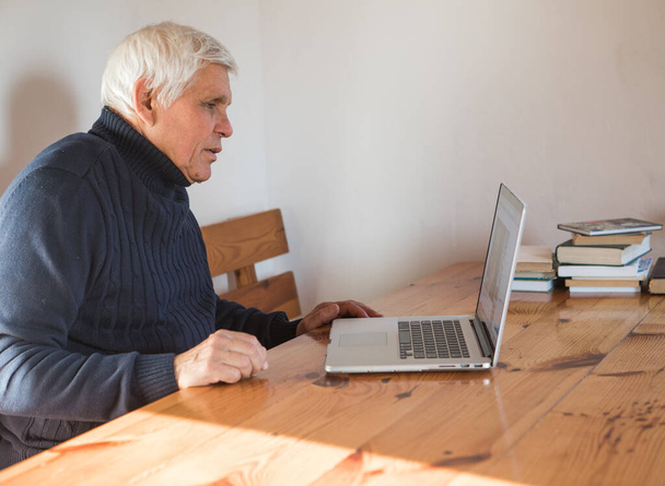 Gülümseyen yaşlı adam kameraya el sallıyor dizüstü bilgisayarında video görüşmesi yapıyor, mutlu yaşlı erkek evde oturup modern teknolojileri ve kablosuz bağlantıyı kullanıyor. - Fotoğraf, Görsel