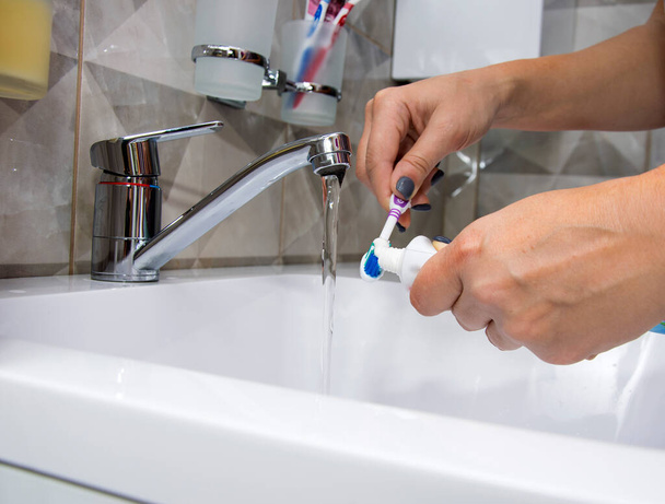 A woman's hand holding a toothbrush and toothpaste under running water from the tap - 写真・画像