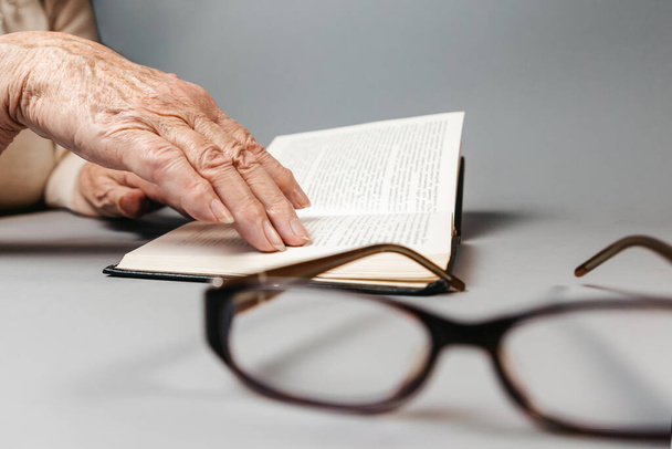 Eine ältere Frau mit faltigen Händen liest ein Buch. Im Vordergrund defokussiert sich die Brille. Hände aus nächster Nähe. Vision-Check und Lesekonzept. - Foto, Bild
