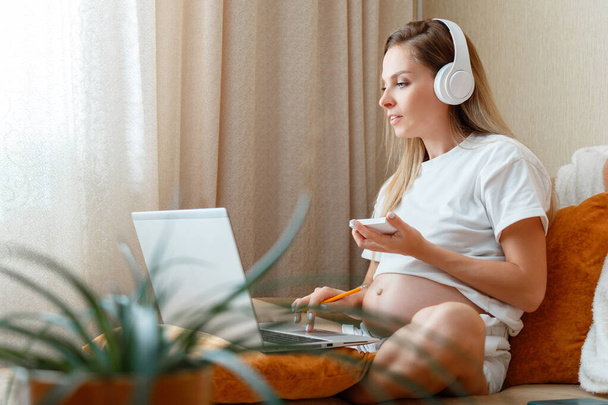 Busy Young pregnant woman freelancer with belly works remotely from home Using laptop headphones. Businesswoman in maternity leave do paperwork. Caucasian 30 s blond woman work at Home office on sofa - Foto, afbeelding