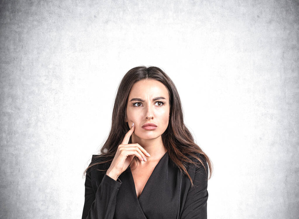 Office woman in black dress with confused, serious look on grey background. Boss making up plans and new ideas. Concept of brainstorm. Copy space blank wall - 写真・画像