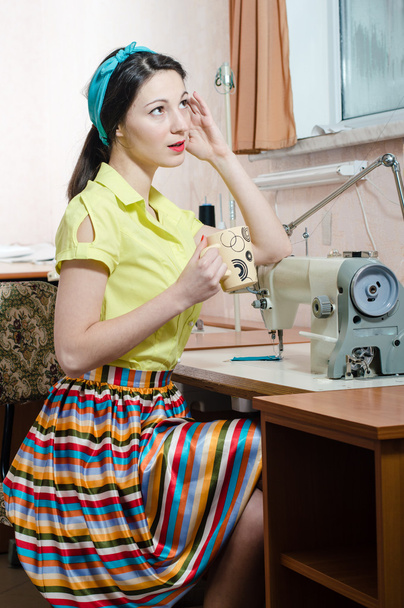 sewing in workshop beautiful brunette young woman pinup girl with red lips and nails in yellow dress blue ribbon on her head holding cup drinking tea & looking at window closeup portrait image - Fotoğraf, Görsel