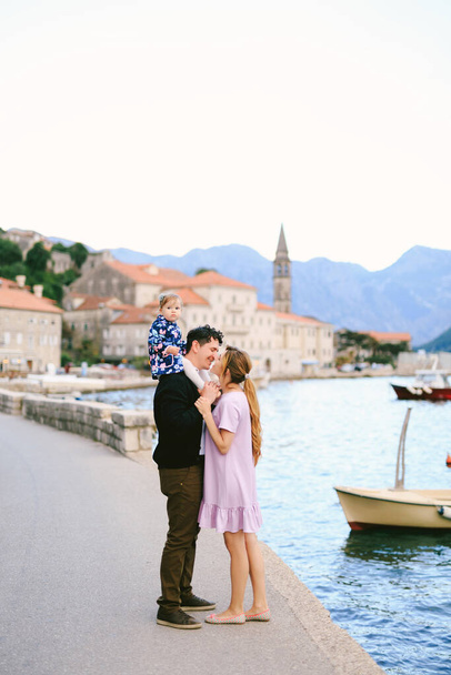 Mom almost kisses smiling dad with daughter on his shoulders on the coast against the background of the sea, boats and ancient buildings of the town of Perast - Photo, Image