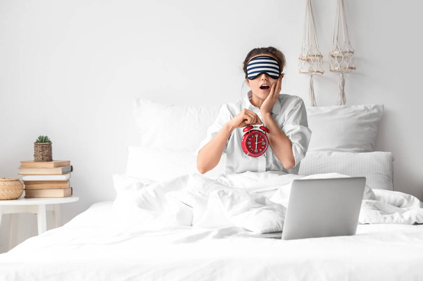 Surprised woman with sleeping mask, alarm clock and laptop sitting in bed - Photo, Image