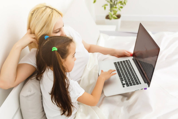 Hymyilevä äiti ja tytär käyttävät kannettavaa tietokonetta sängyssä kotona
 - Valokuva, kuva