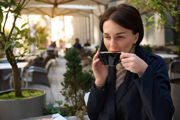 魅力的な若い成功したブルネットの女性はコーヒーを飲み、美しい晴れた日にコーヒーブレイク中に屋外でリラックスしながらスマートフォンで話しています - 写真・画像