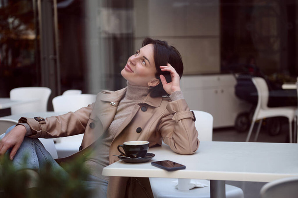 Attrayant élégant jeune femme brune à la mode en tenue décontractée regarde reposer sur la terrasse d'été de la cafétéria d'été profiter du temps chaud et ensoleillé de l'automne pendant la pause café - Photo, image