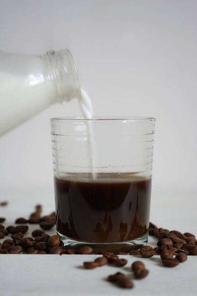                  Il latte di una bottiglia di vetro viene versato in una tazza di vetro con caffè. Chicchi di caffè e caffè in un bicchiere su sfondo bianco.               - Foto, immagini