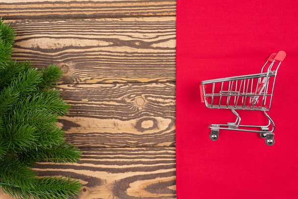Small trolley, mini shopping cart on red textile, Christmas tree branch on wooden background. New year fair, market, sale concept with copy space - Photo, Image