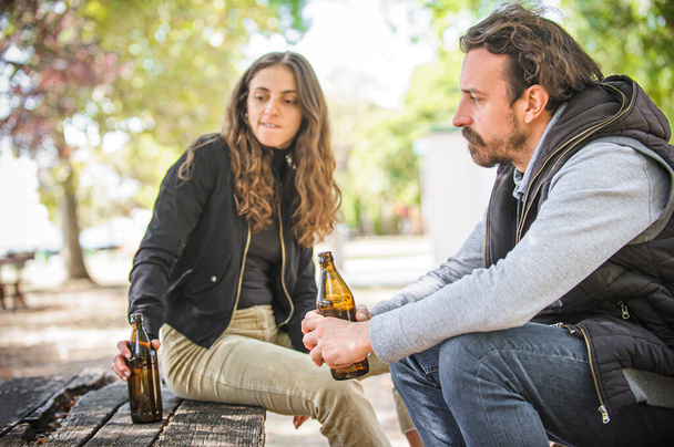 Young couple in quarrel conflict, boyfriend and girlfriend drinks beer from bottle on wooden bench with table in forest park outdoor. Real life scene. Relationship disagree concept. - Valokuva, kuva