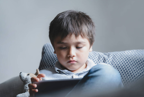 Portré gyerek ül a kanapén néz rajzfilm tabletta, Aranyos fiú játszik játék touch pad, Cinematikus beltéri portré Gyermek pihentető egyedül a nappaliban, Új normális életmód - Fotó, kép