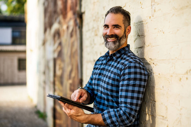 Portret współczesnego biznesmena z brodą za pomocą tabletu cyfrowego stojącego przed murem murowanym na zewnątrz. - Zdjęcie, obraz