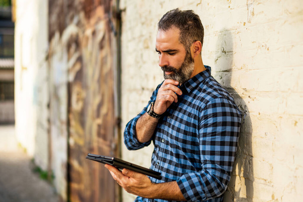 Portrait of modern businessman  with beard thinking and  using digital tablet while  standing in front of brick wall  outdoor. - 写真・画像