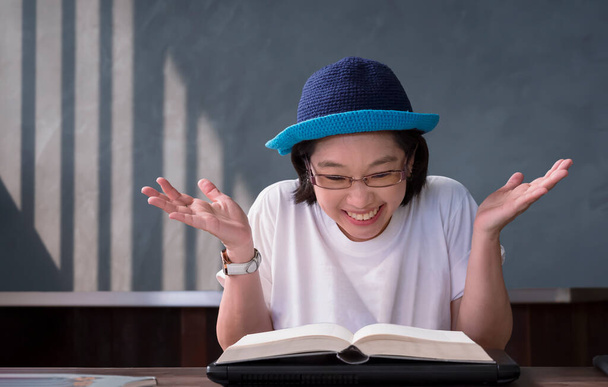アジア系女子高生のポートレートは、決勝試験後の教科書で内容が正しいことを確認しながら、興奮して笑顔とオープンパームジェスチャーを示す - 写真・画像