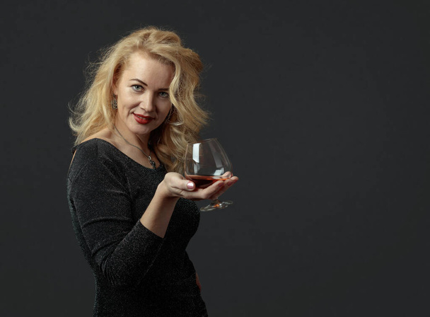 An attractive mature woman in a black evening dress is tasting cognac. Beautiful forty years blonde with long hair on a dark background. Copy space. - Foto, Imagen