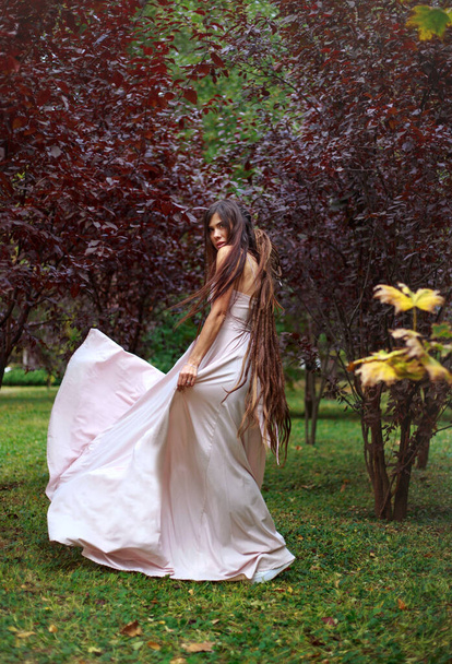 Mujer joven con un largo vestido rosa corre en un baile sobre la hierba verde, en el jardín en un parque de árboles rojos - Foto, Imagen