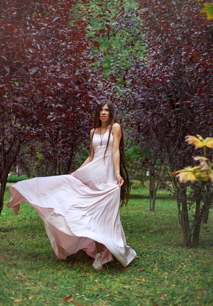Молода жінка в довгій рожевій сукні біжить в танці на зеленій траві, в саду в парку червоних дерев
 - Фото, зображення