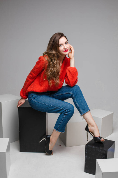 Studio shot of attractive brunette with red lips in stylish casual red pullover and denim jeans and black heels sitting on black and white cubes smiling at camera. - Photo, image