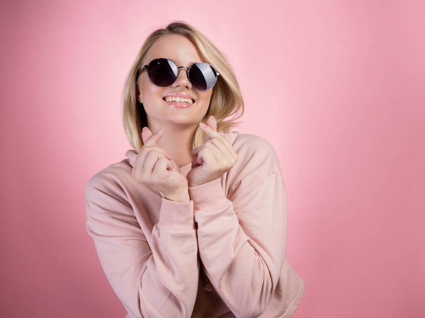 A cheerful young blonde in sunglasses makes a sign of love, shows a heart with her fingers. - Fotoğraf, Görsel