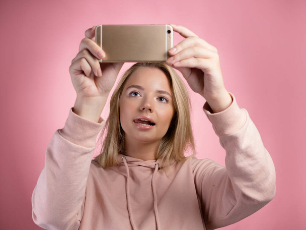 An attractive young woman in a pink hoodie uses a smartphone for mobile games. - Fotó, kép
