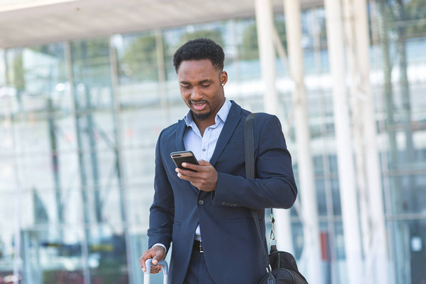successful young confident African American businessman moving with cellphone in hands against modern office building background downtown black business man walks using a mobile phone in formal suit - Photo, image
