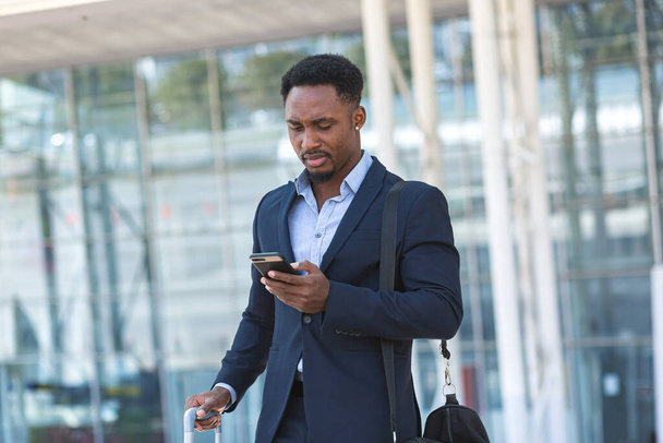 successful young confident African American businessman moving with cellphone in hands against modern office building background downtown black business man walks using a mobile phone in formal suit - 写真・画像