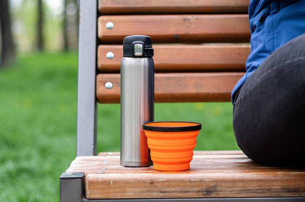 Termo de acero y taza de silicona. Platos turísticos en el banco. Beber café en el parque. Beber té en el parque. - Foto, imagen