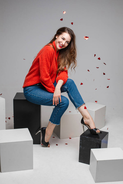 Joyful smiling young girl under festive confetti wearing red sweater, jeans posing on black and white cubes in studio - Photo, image