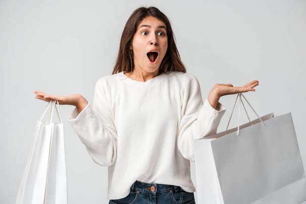 Überraschte Frau in lässigem Pullover mit Einkaufstüten, mit offenem Mund und schockiertem Gesichtsausdruck. Indoor-Studio isoliert auf weißem Hintergrund aufgenommen  - Foto, Bild