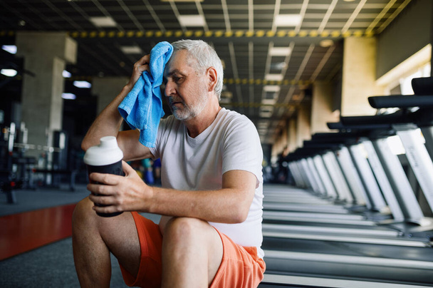Vieil homme fatigué avec serviette boit de l'eau dans la salle de gym - Photo, image