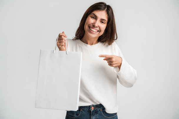 Šťastná veselá žena ukazující prstem na papírovou nákupní tašku v ruce, spokojená s nákupy, velký výprodej. Vnitřní studio záběr izolované na bílém pozadí  - Fotografie, Obrázek