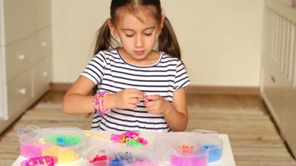 Una niña de cinco años se dedica a tejer accesorios de bandas elásticas. Un conjunto de herramientas de tejido y un montón de caucho colorido en recipientes de plástico en la mesa delante del niño. Pasatiempos para niños - Imágenes, Vídeo